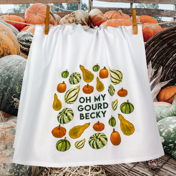 Oh My Gourd Becky dishtowel shown on a clothesline with pumpkins and gourds in the background.