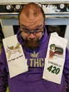 A customer submitted photo of a man sitting in front of his oven with the I'm high as fuck dishtowel over one shoulder and the prebake to 420 dishtowel over the other