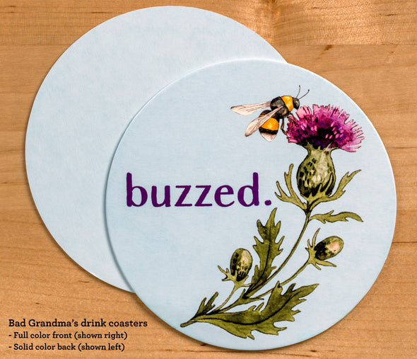 The front and back of the round drink coaster. The front features the text Buzzed and a watercolor-style bee and thistle illustration. The back of the coaster is baby blue, which matches the background on the front of the coaster.