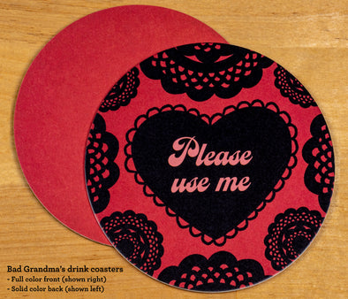 The front and back of the round drink coaster. The front features the text Please Use Me and has illustrations of a black heart shape with a lace edge and doilies, the background is red. The back is solid red. 