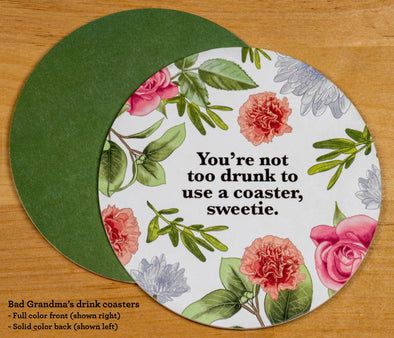 The front and back of the round drink coaster. The front features the text You're not too drunk to use a coaster, sweetie in black font on a white background, surrounded with illustrations of flowers and leaves. The leaves are green and the flowers are pink and orange. The back is solid green. 