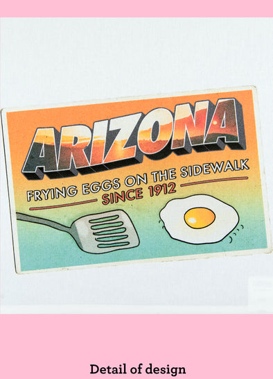 Close-up of a dishtowel with the phrase Arizona, Frying Eggs on the Sidewalk and an illustration of a postcard with a fried egg and a spatula.