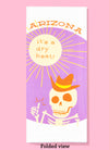 Folded dishtowel with the phrase Arizona, It's a Dry Heat and an illustration of a skeleton giving a thumbs-up gesture underneath a summer sun.