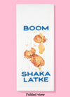 Folded dishtowel with illustration of potato latkes, sour cream, and apple sauce and the text Boom Shaka Latke.