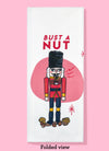 Folded dishtowel with the phrase Bust a Nut with an illustration of a wooden nutcracker standing among unshelled nuts.