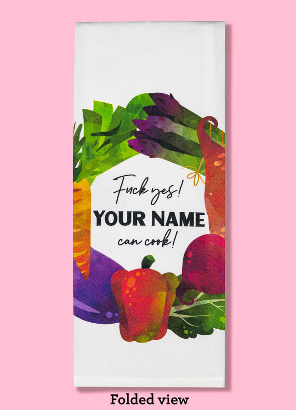 Folded dishtowel demonstrating how this product can be personalized. It features text that says Fuck Yes Your Name Can Cook and an illustration of wreath of vegetables.