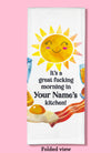 Folded dishtowel demonstrating how this product can be personalized. It features text that says It's a Great Fucking Morning In Your Name's Kitchen and an illustration of a smiling sun and breakfast foods.