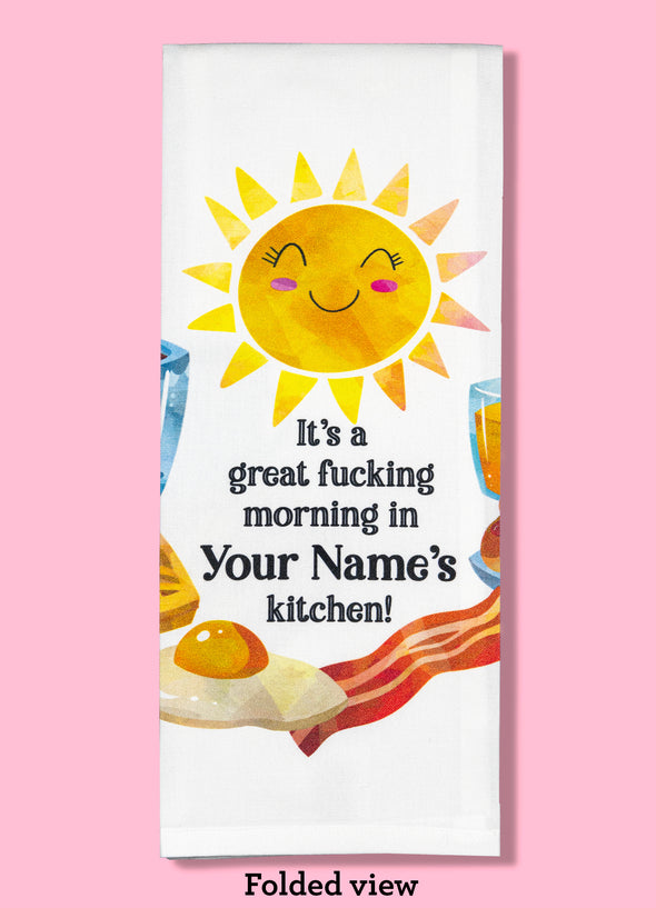 Folded dishtowel demonstrating how this product can be personalized. It features text that says It's a Great Fucking Morning In Your Name's Kitchen and an illustration of a smiling sun and breakfast foods.