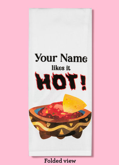 Folded dishtowel demonstrating how this product can be personalized. It features text that says Your Name likes it hot and an illustration of a tortilla chip in a bowl of salsa.