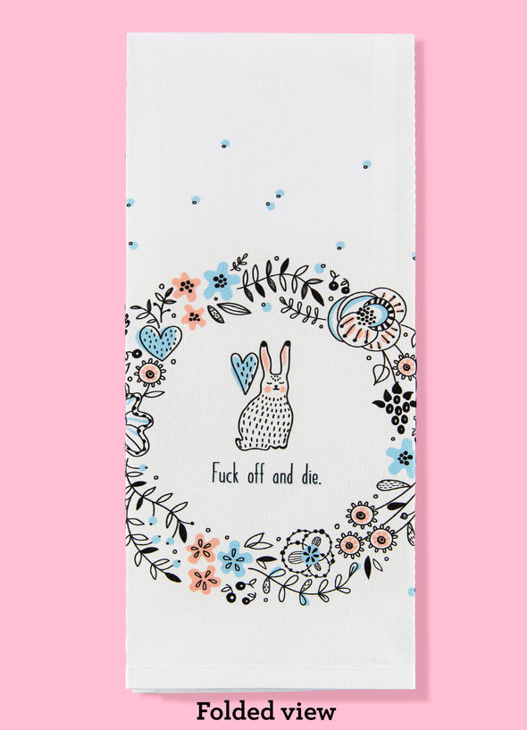 Folded dishtowel with an illustration of a wreath of flowers and a bunny and the phrase Fuck Off and Die.