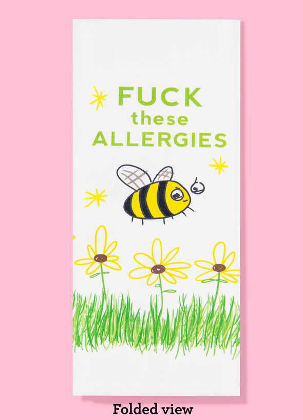 Folded dishtowel with an illustration of a bee, grass and flowers with the phrase Fuck These Allergies.