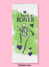 Folded dishtowel with an illustration of a winking skeleton holding a femur bone surrounded by purple hearts and the phrase I Have a Boner.