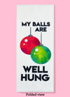Folded dishtowel with the phrase my balls are well hung. The illustration is of two Christmas ornament balls, one red and one green