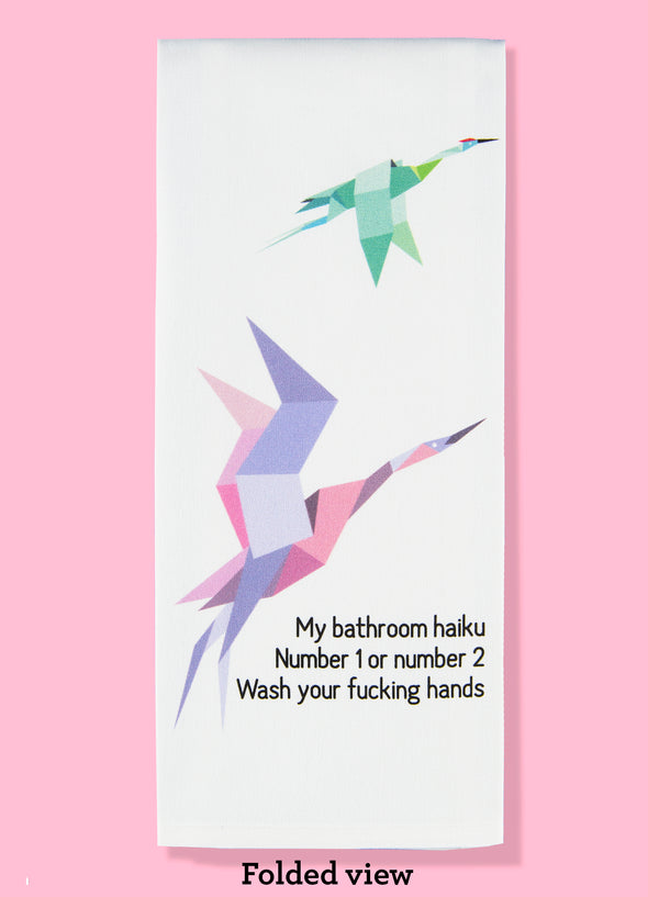 Folded dishtowel with the haiku: my bathroom haiku, number 1 or number 2, wash your fucking hands. The illustration is of flying paper cranes