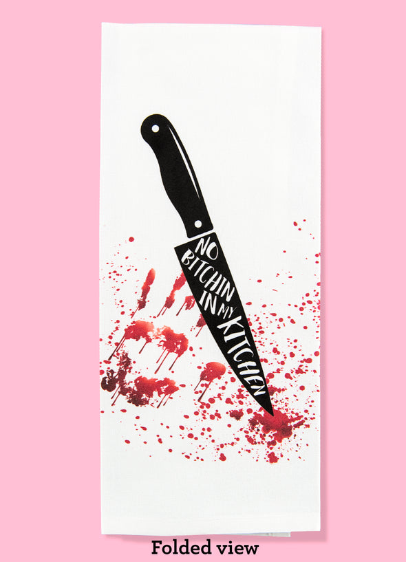 Folded dishtowel with an illustration of a giant black knife with the phrase no bitchin' in my kitchen on the knife. In the background is an illustration of a blood splatter and a bloody hand print