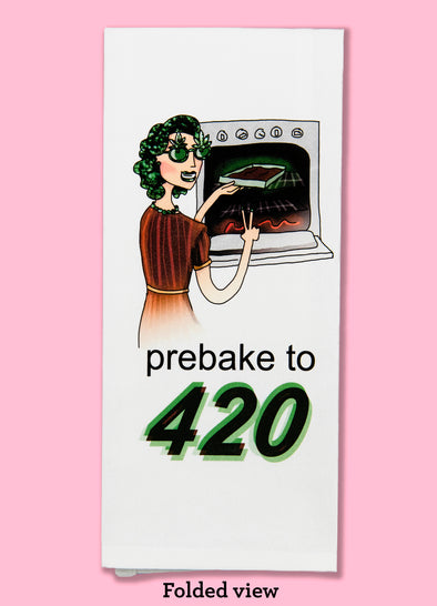 Folded dishtowel with the phrase prebake to 420. The illustration is of a cartoon woman pulling brownies out of the ovens, holding up a peace sign, and wearing green sunglasses with marijuana leaves on them