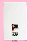 Unfolded dishtowel with the phrase prebake to 420. The illustration is of a cartoon woman pulling brownies out of the ovens, holding up a peace sign, and wearing green sunglasses with marijuana leaves on them