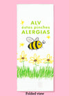 Folded dishtowel with the phrase ALV éstas pinches alergias and an illustration of a bee, flowers, and grass.