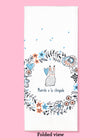 Folded dishtowel with an illustration of a wreath of flowers and a bunny and the phrase muerete ala chingada