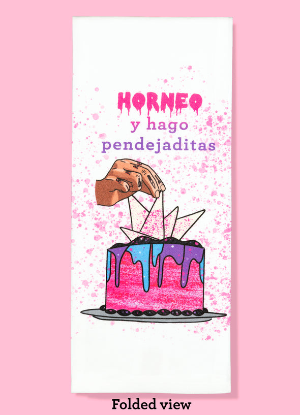 Folded dishtowel with an illustration of a tattooed hand decorating a cake and the Spanish phrase Horneo y hago pendejaditas.
