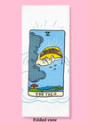 Folded dishtowel of an illustration of a faux tarot card featuring a giant hand emerging from a dark cloud holding a fully loaded taco above a coastal village with the phrase The Taco