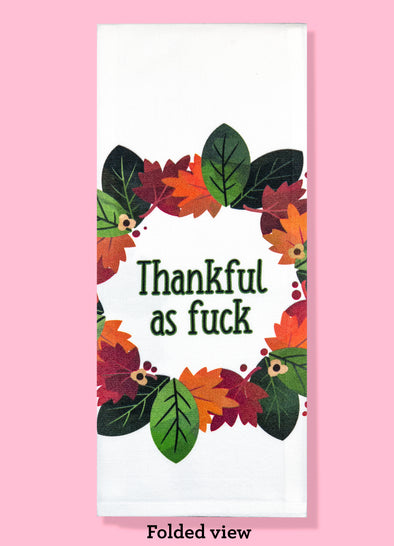 Folded dishtowel with the phrase thankful as fuck. The text is surrounded by a circle of illustrations of autumn leaves