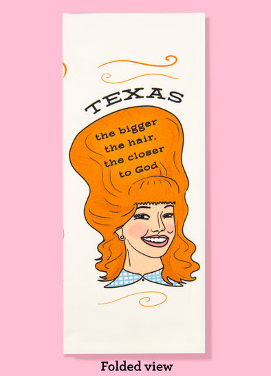 Folded dishtowel with the phrase Texas the bigger the hair, the closer to God. The illustration is of a cartoon smiling woman with large red hair.