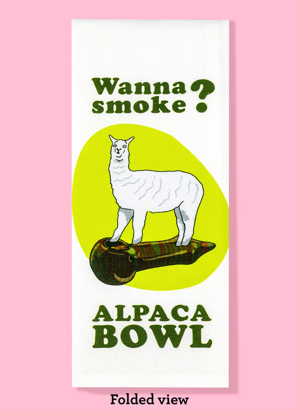 Folded dishtowel with the humorous phrase Wanna smoke? alpaca bowl. The text is in a cartoon illustration of a minature alpaca standing on top of a marijuana pipe.