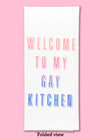 Folded dishtowel with the phrase welcome to my gay kitchen. The text fades from a peach color to blue.