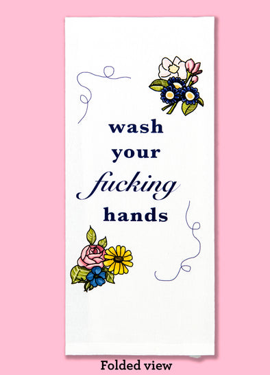 Folded dishtowel with the phrase wash your fucking hands. The text is surrounded by illustrations of flowers in the upper right and lower left of the design and dark blue squiggles in the upper left and lower right