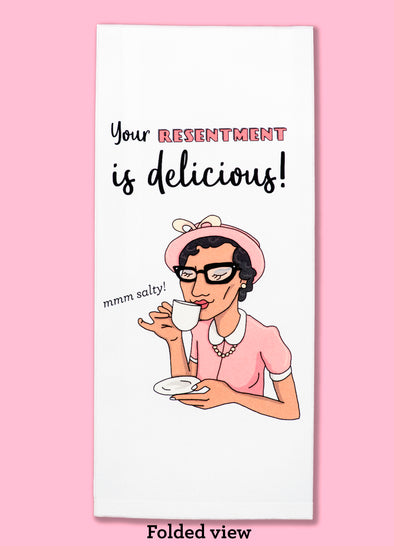 Folded dishtowel with the phrase Your Resentment is Delicious!. The illustration shows a woman sipping tea with an illustration of her saying mmm salty!.